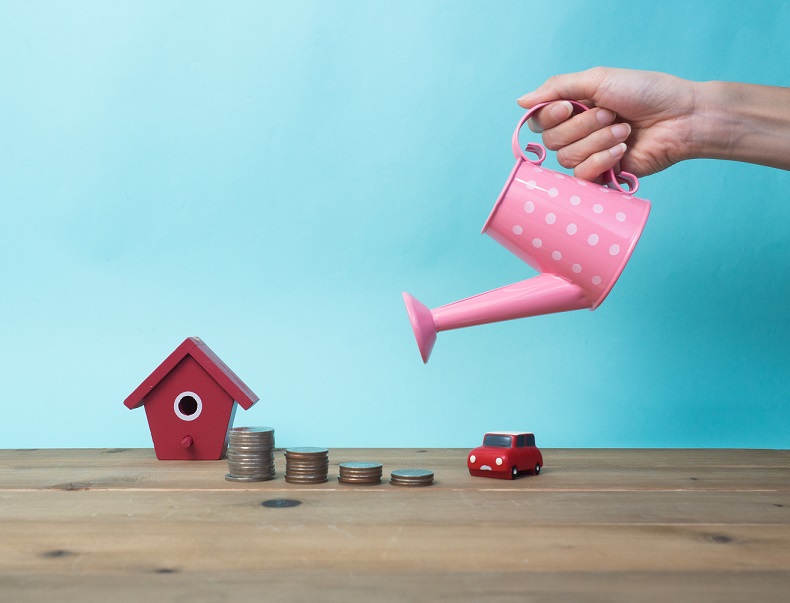 hand holding watering can over mini house and stack of coins